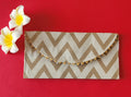 A V-shaped red and gold shagun envelope with a zigzag pattern and decorative golden trimmings, displayed on an orange background with a frangipani flower beside it.