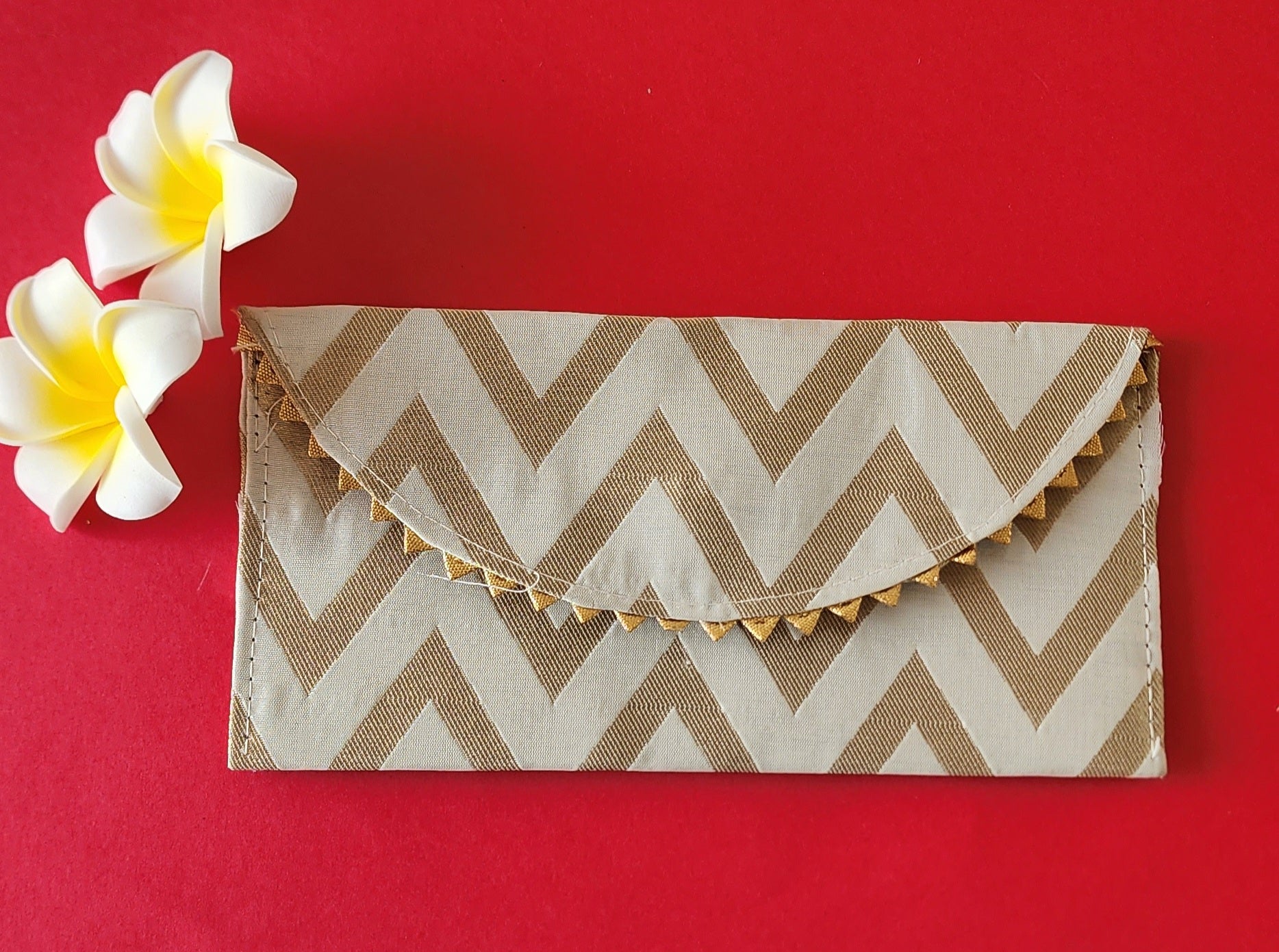 A V-shaped red and gold shagun envelope with a zigzag pattern and decorative golden trimmings, displayed on an orange background with a frangipani flower beside it.