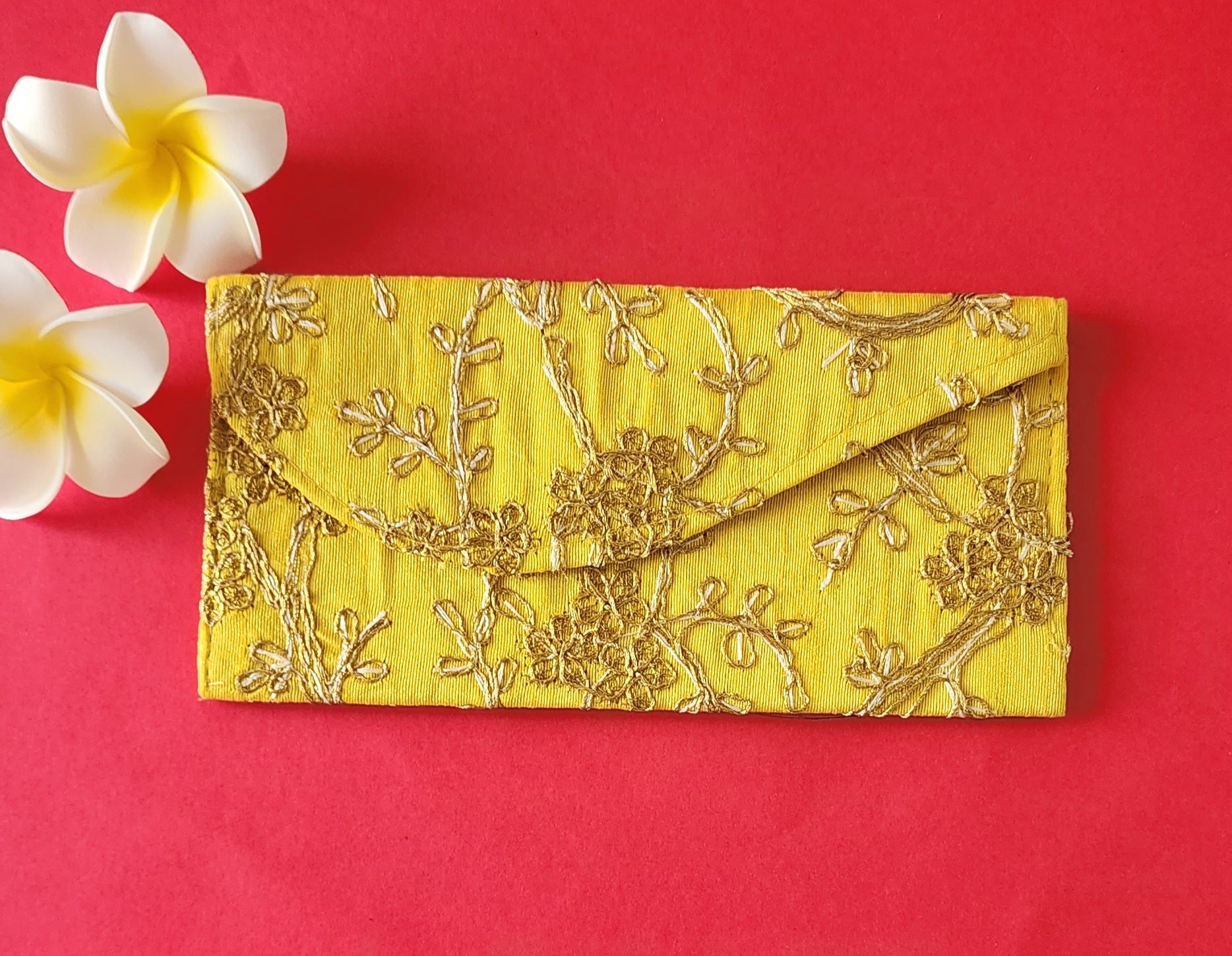 A yellow color envelope with gold floral embroidery, placed on an orange surface with two white flowers at the top left corner.
