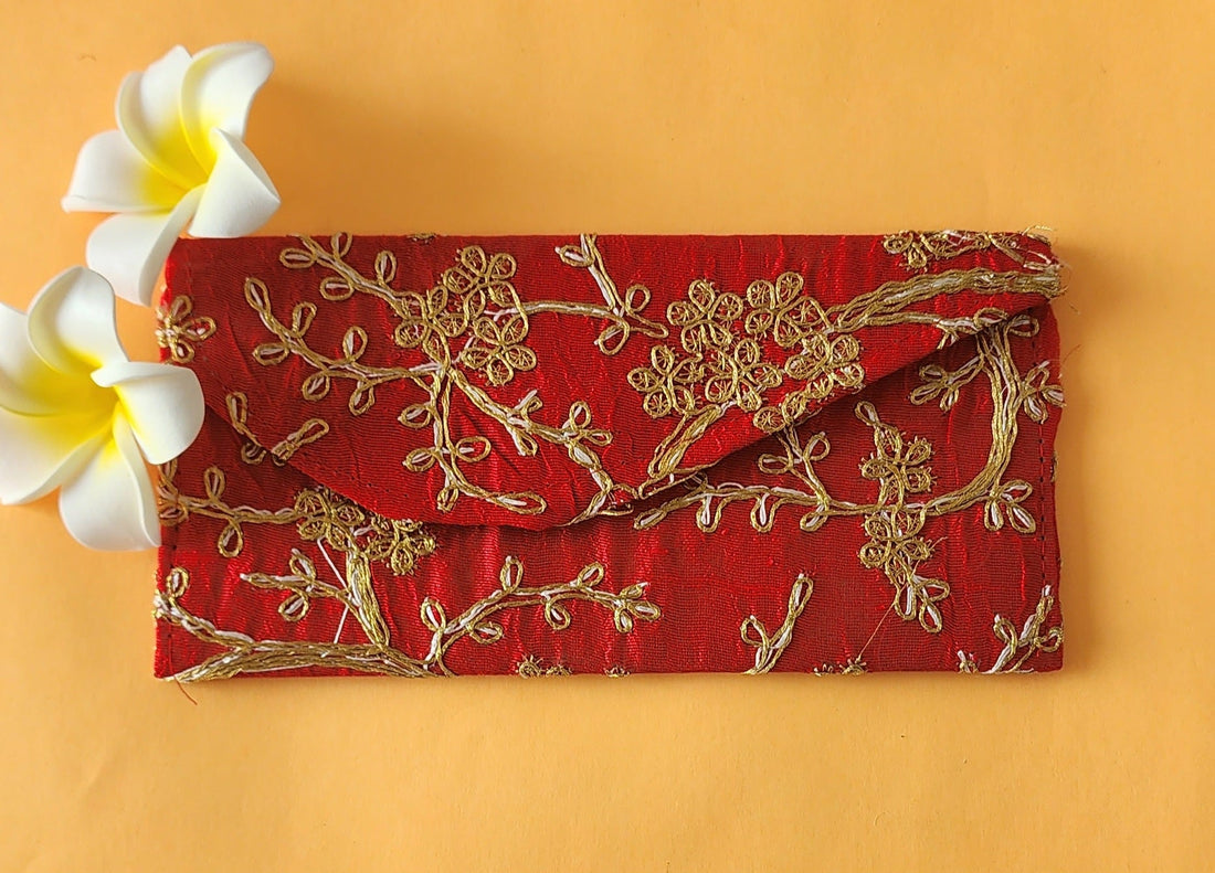 A red envelope with gold floral embroidery, placed on an orange surface with two white flowers at the top left corner.