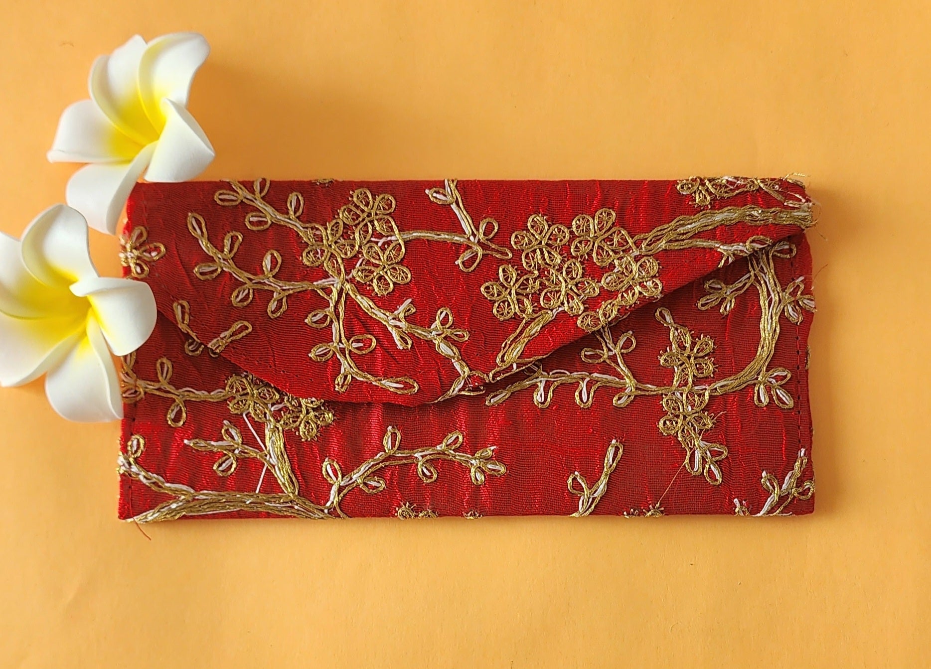 A red envelope with gold floral embroidery, placed on an orange surface with two white flowers at the top left corner.
