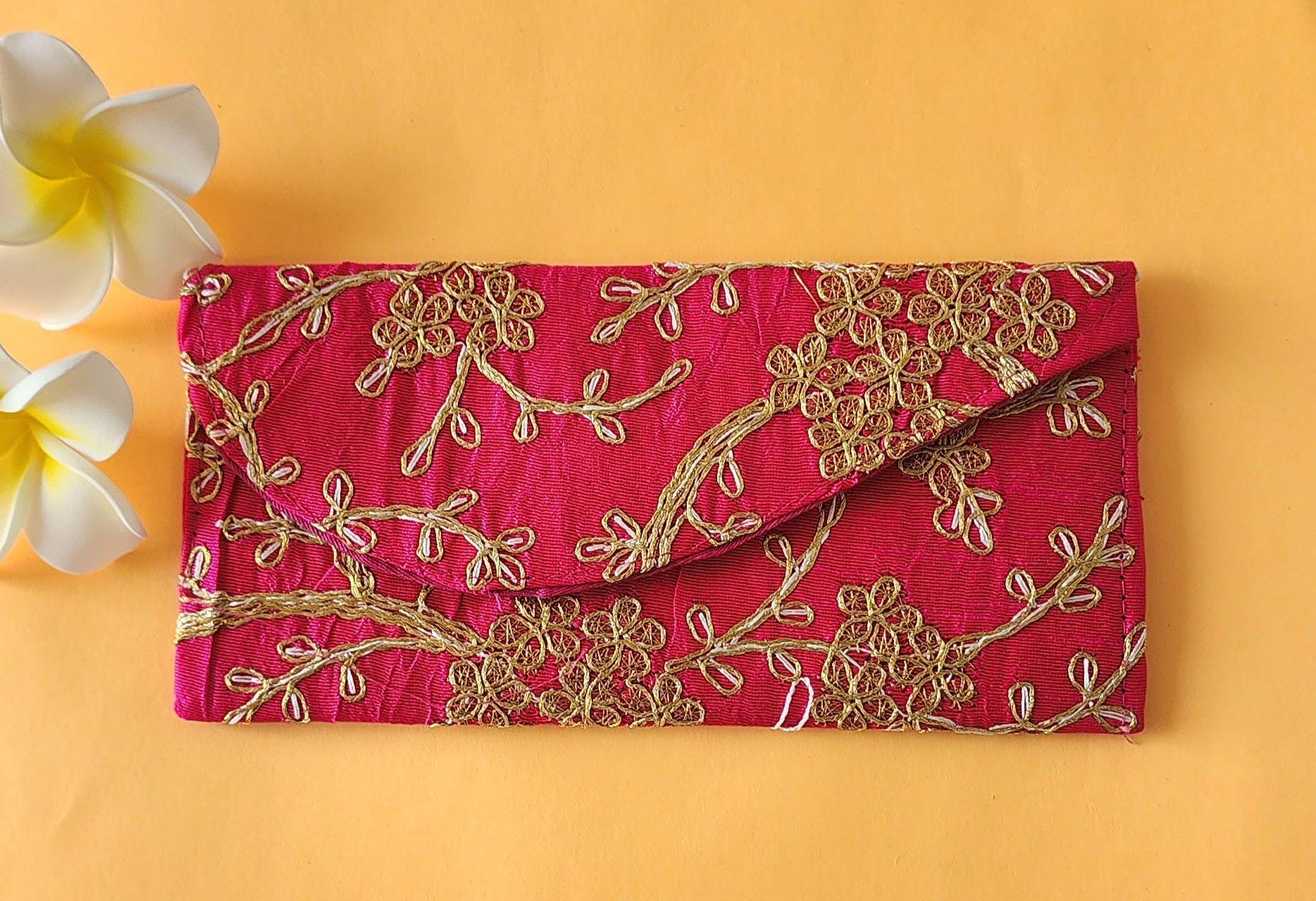 A dark Pink envelope with gold floral embroidery, placed on an orange surface with two white flowers at the top left corner.