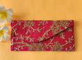 A red envelope with gold floral embroidery, placed on an orange surface with two white flowers at the top left corner.