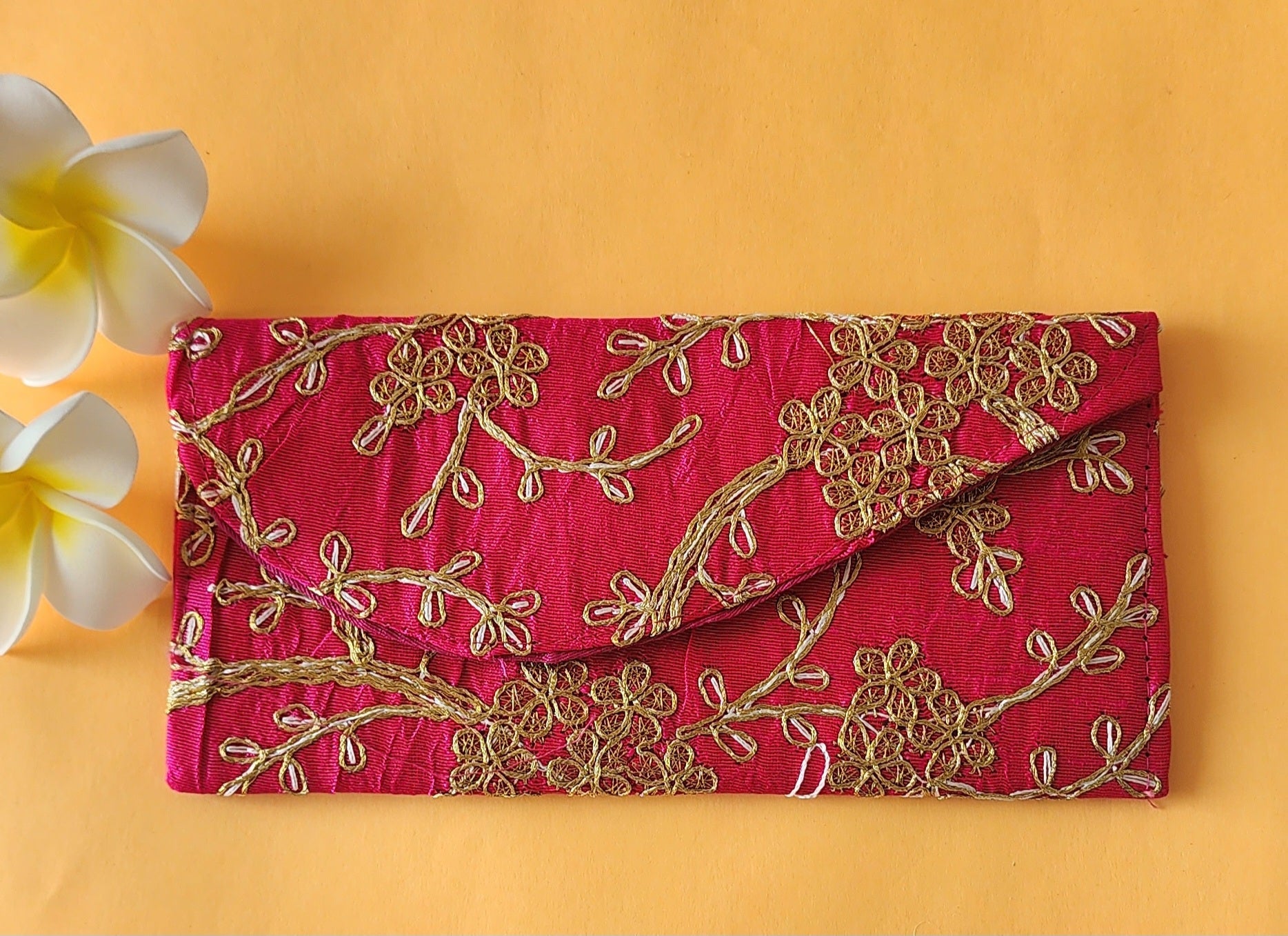 A red envelope with gold floral embroidery, placed on an orange surface with two white flowers at the top left corner.
