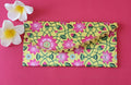 A multicolored floral patterned Designer Shagun Envelope placed on a pink surface with two white flowers at the top left corner.