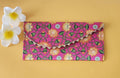 A multicolored floral patterned Designer Shagun Envelope placed on a pink surface with two white flowers at the top left corner.
