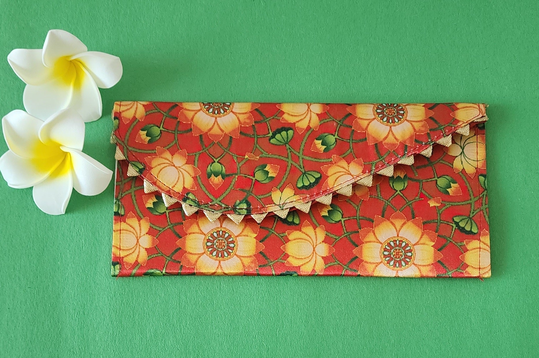 A multicolored floral patterned Designer Shagun Envelope placed on a pink surface with two white flowers at the top left corner.