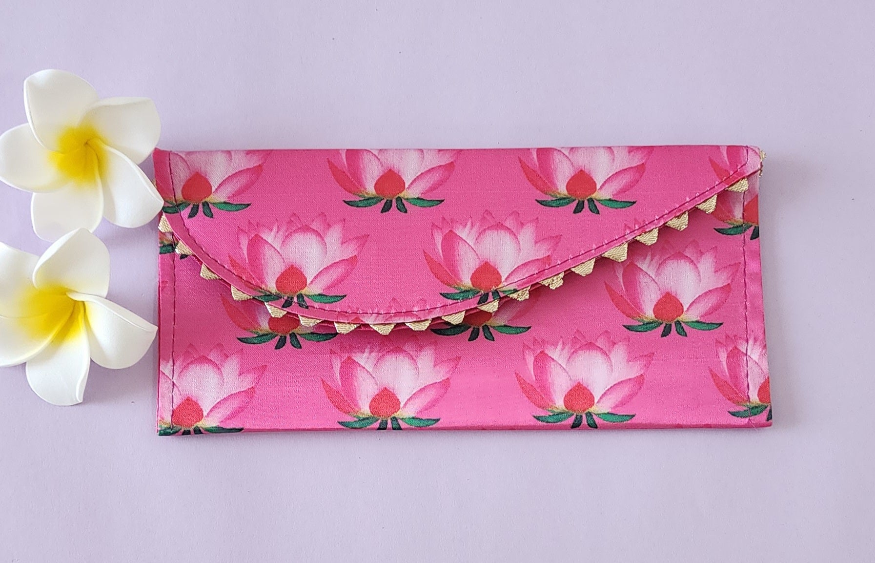 A multicolored floral patterned Designer Shagun Envelope placed on a pink surface with two white flowers at the top left corner.