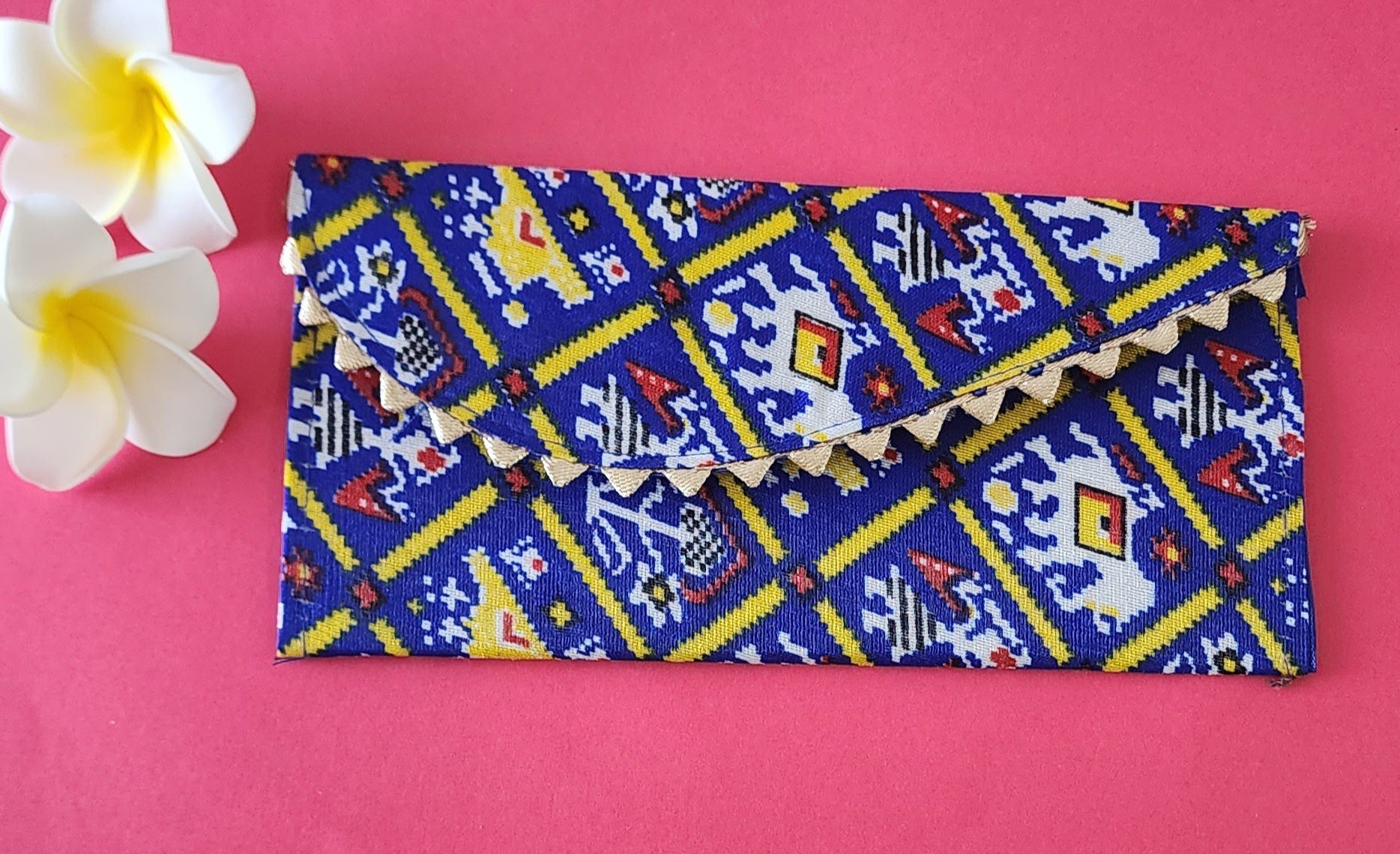 A multicolored floral patterned Designer Shagun Envelope placed on a pink surface with two white flowers at the top left corner.