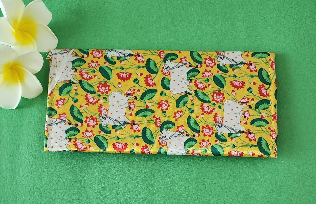 A yellow shagun envelope featuring a traditional Pichwai art-style design of a white Kamdhenu cow surrounded by red lotus flowers and green leaves, with a decorative scalloped edge, placed on a green surface alongside two white and yellow frangipani flowers.