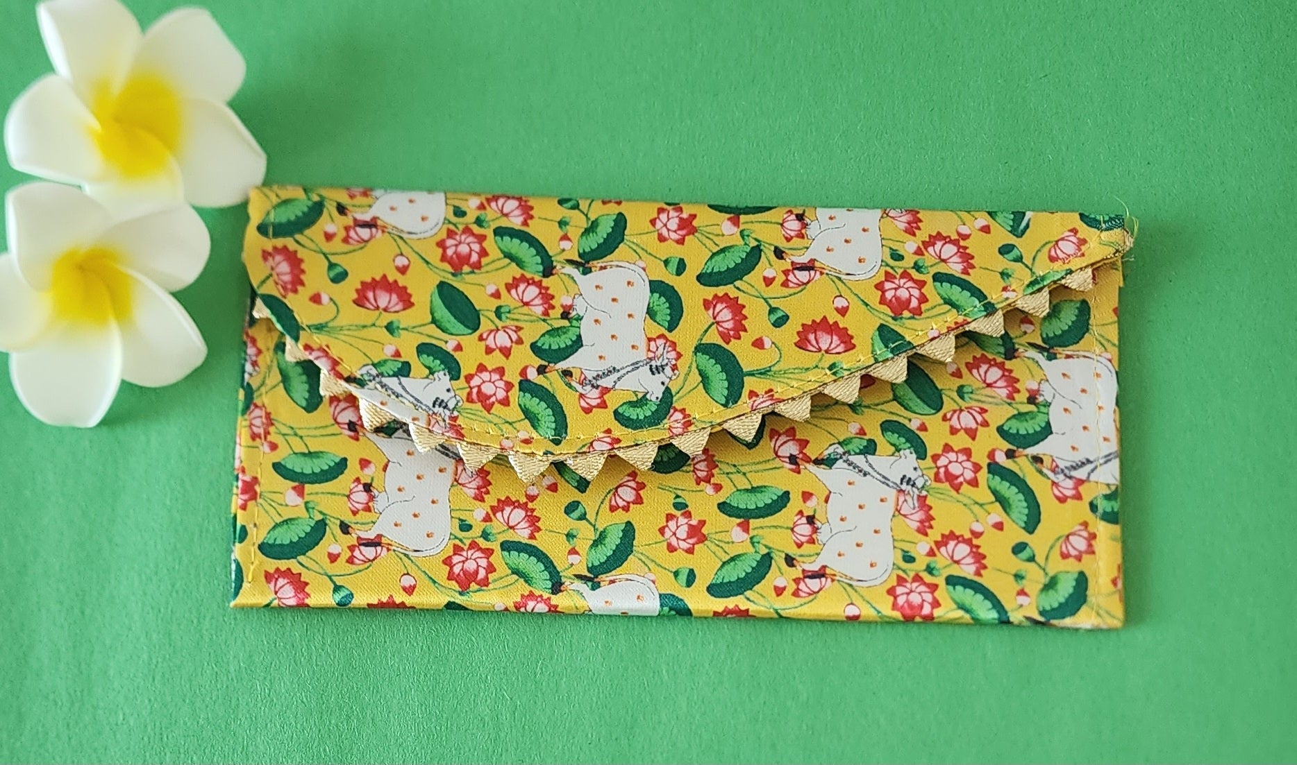 A yellow shagun envelope featuring a traditional Pichwai art-style design of a white Kamdhenu cow surrounded by red lotus flowers and green leaves, with a decorative scalloped edge, placed on a green surface alongside two white and yellow frangipani flowers.