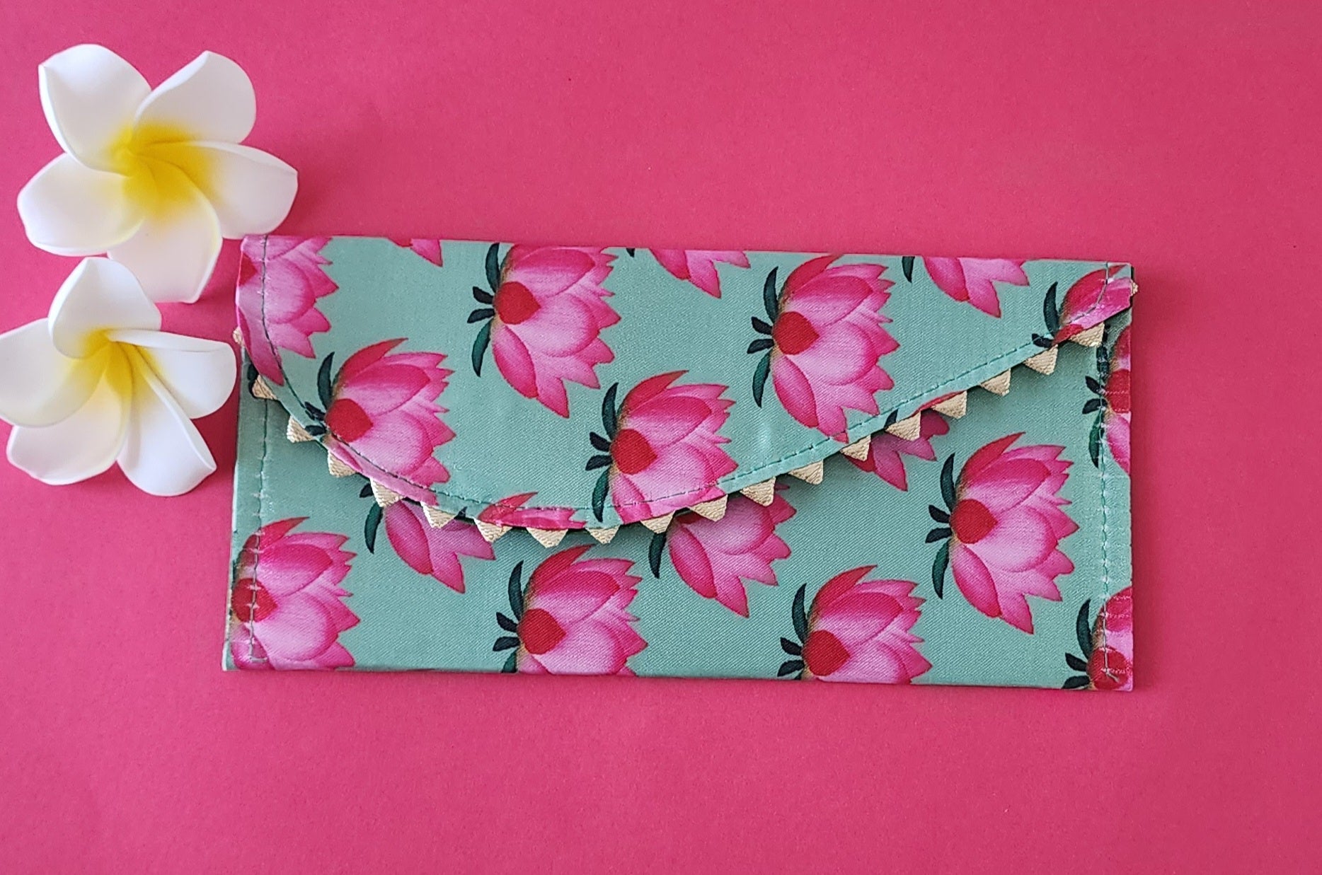 A multicolored floral patterned Designer Shagun Envelope placed on a pink surface with two white flowers at the top left corner.