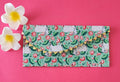 A vibrant green fabric envelope or clutch with a decorative gold trim featuring small tassels. The fabric is patterned with white Kamdhenu cows and pink lotus flowers. Two white plumeria flowers are placed on the left side, all set against a bright pink background.