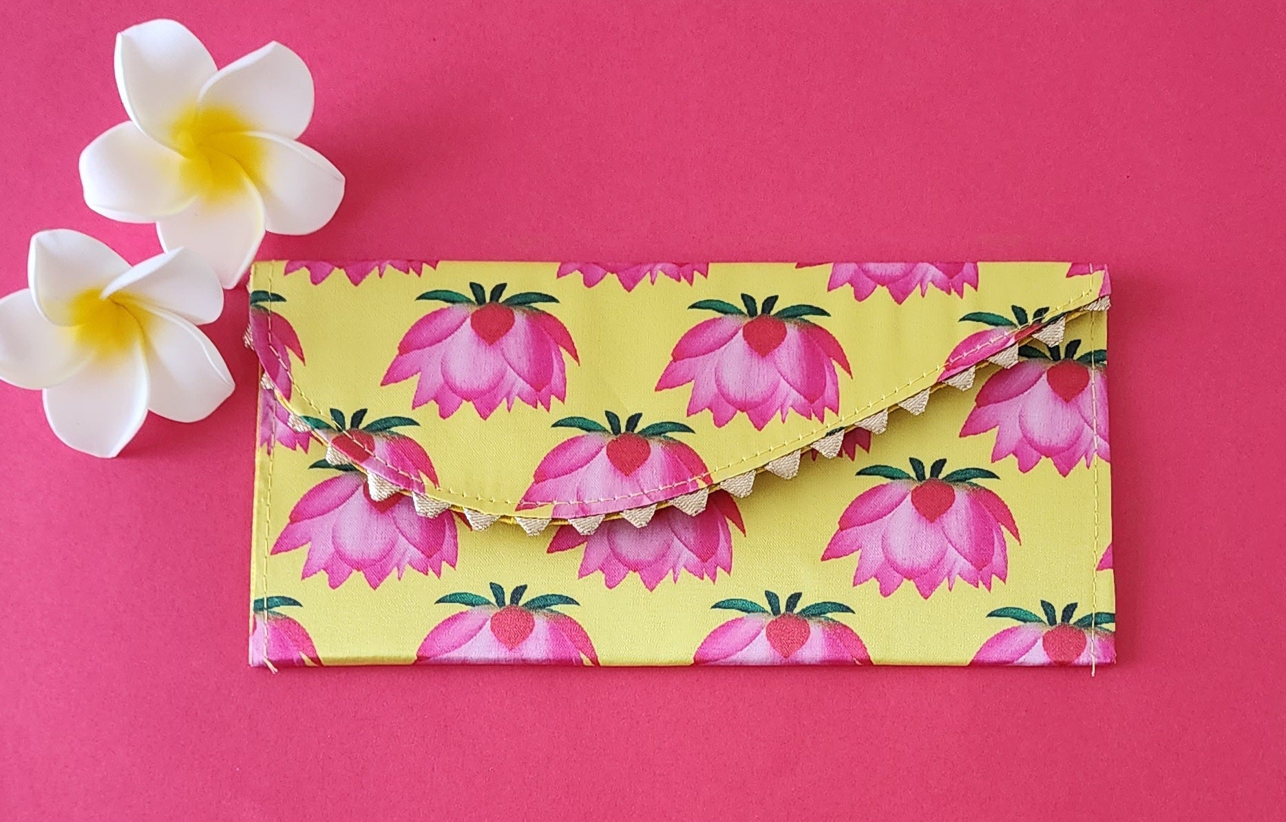 A multicolored floral patterned Designer Shagun Envelope placed on a pink surface with two white flowers at the top left corner.