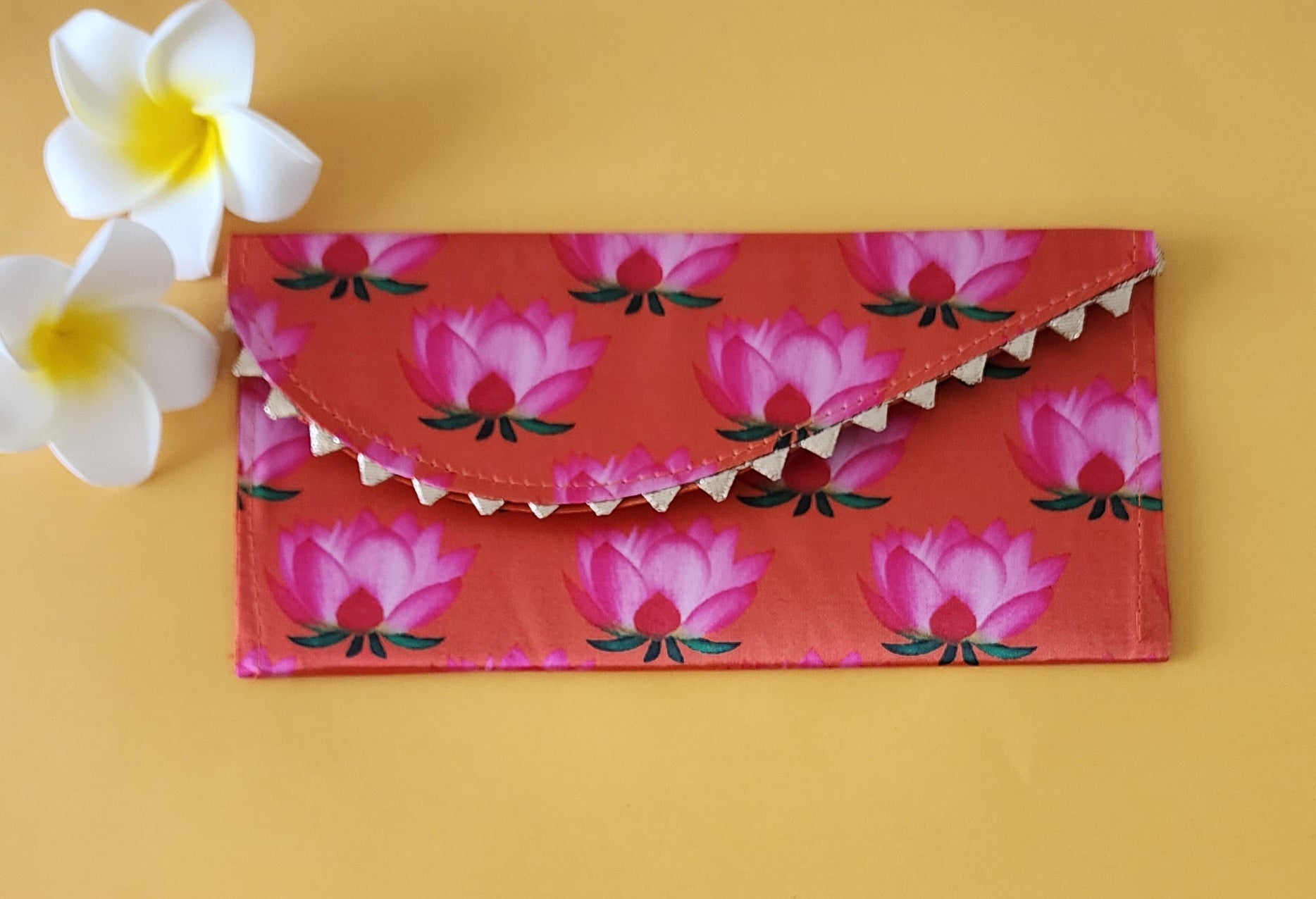 A multicolored floral patterned Designer Shagun Envelope placed on a pink surface with two white flowers at the top left corner.