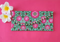 A multicolored floral patterned Designer Shagun Envelope placed on a pink surface with two white flowers at the top left corner.