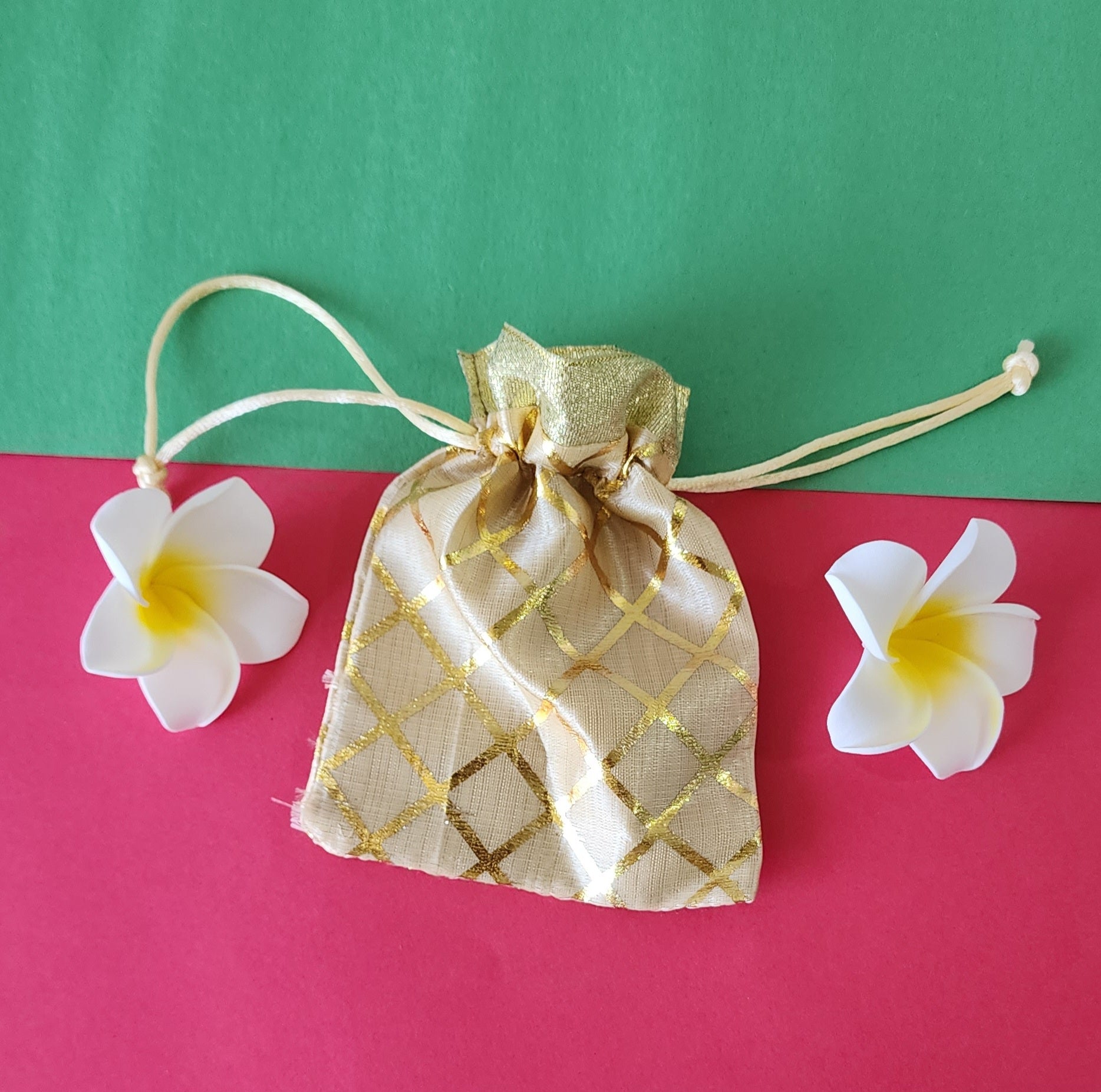 gold patterned fabric potli pouch with a drawstring closure, accompanied by two white flowers on a green background.