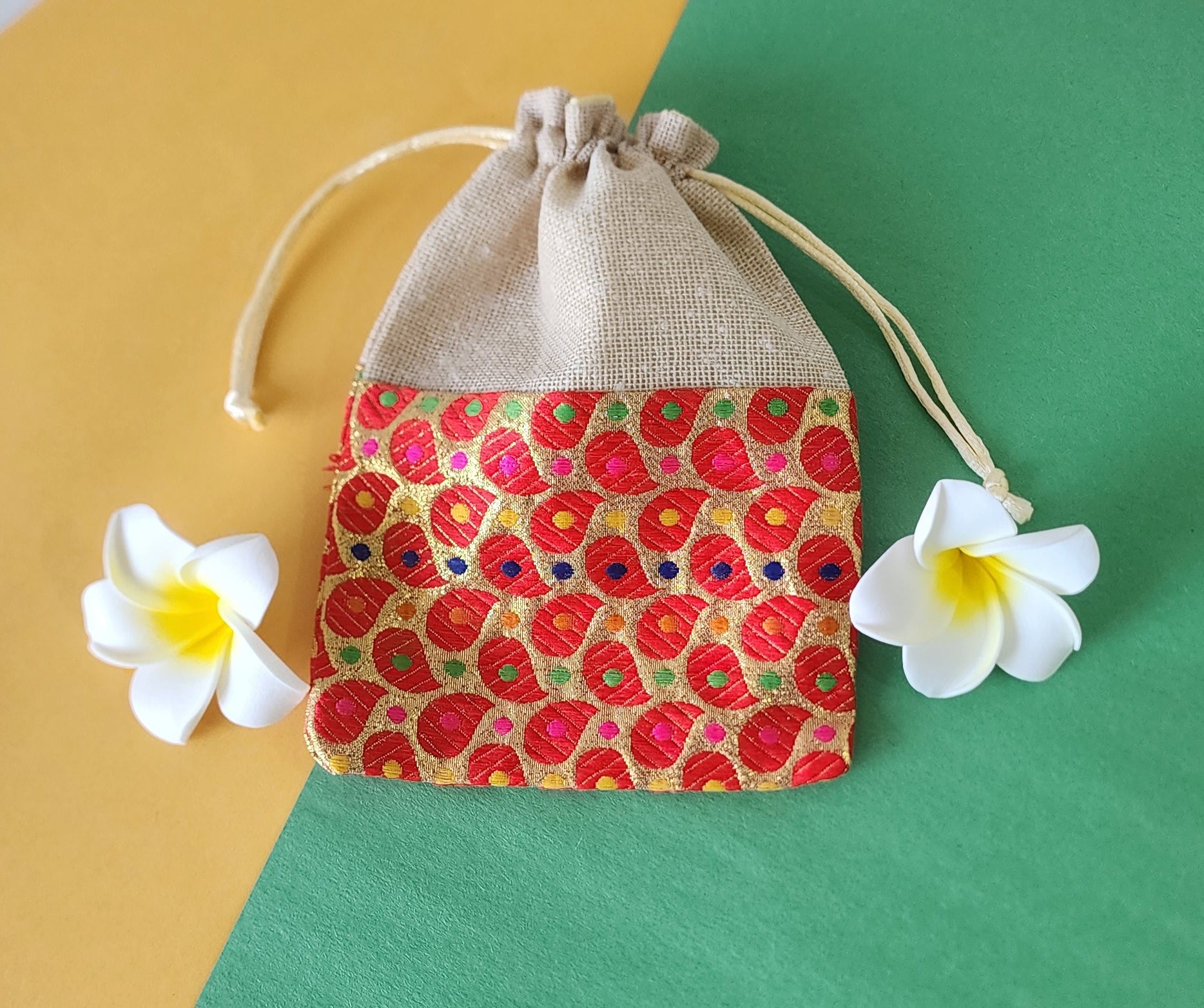 A red gift potli pouch with a floral pattern and drawstring closure, displayed on a flat surface with two white flowers beside it.