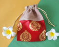A red and gold patterned fabric potli pouch with a drawstring closure, accompanied by two white flowers on a green background.