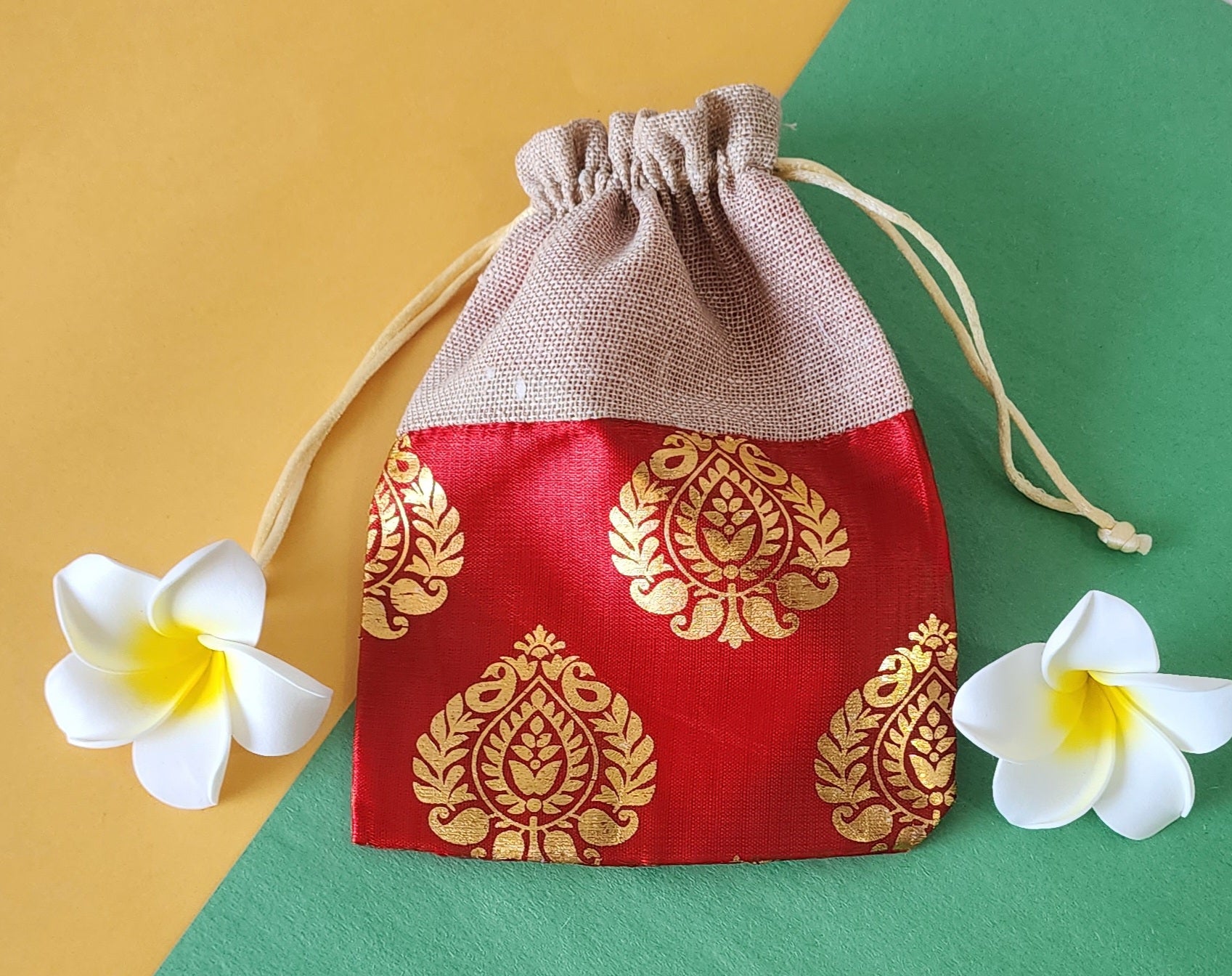 A red and gold patterned fabric potli pouch with a drawstring closure, accompanied by two white flowers on a green background.