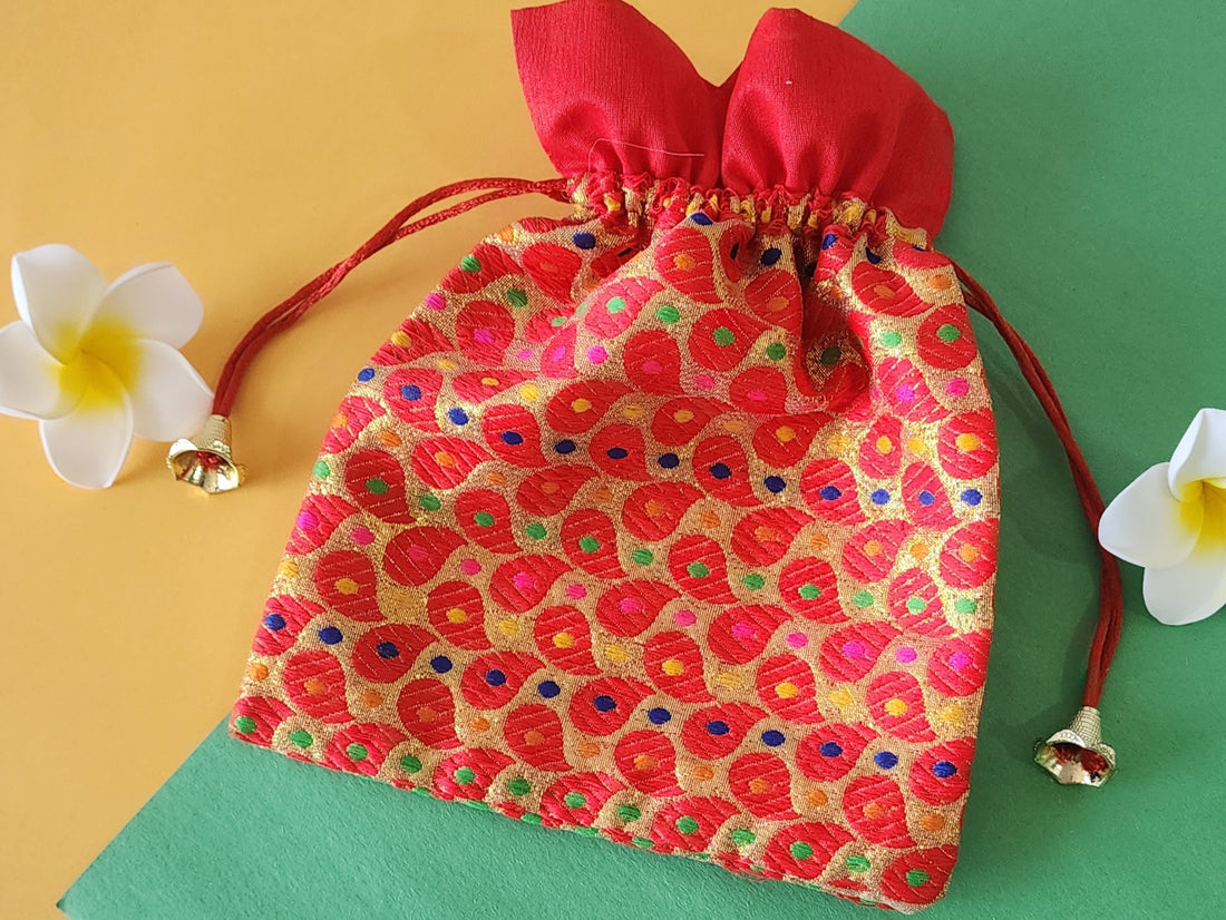 A red gift potli pouch with a floral pattern and drawstring closure, displayed on a flat surface with two white flowers beside it.
