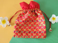 A red gift potli pouch with a floral pattern and drawstring closure, displayed on a flat surface with two white flowers beside it.