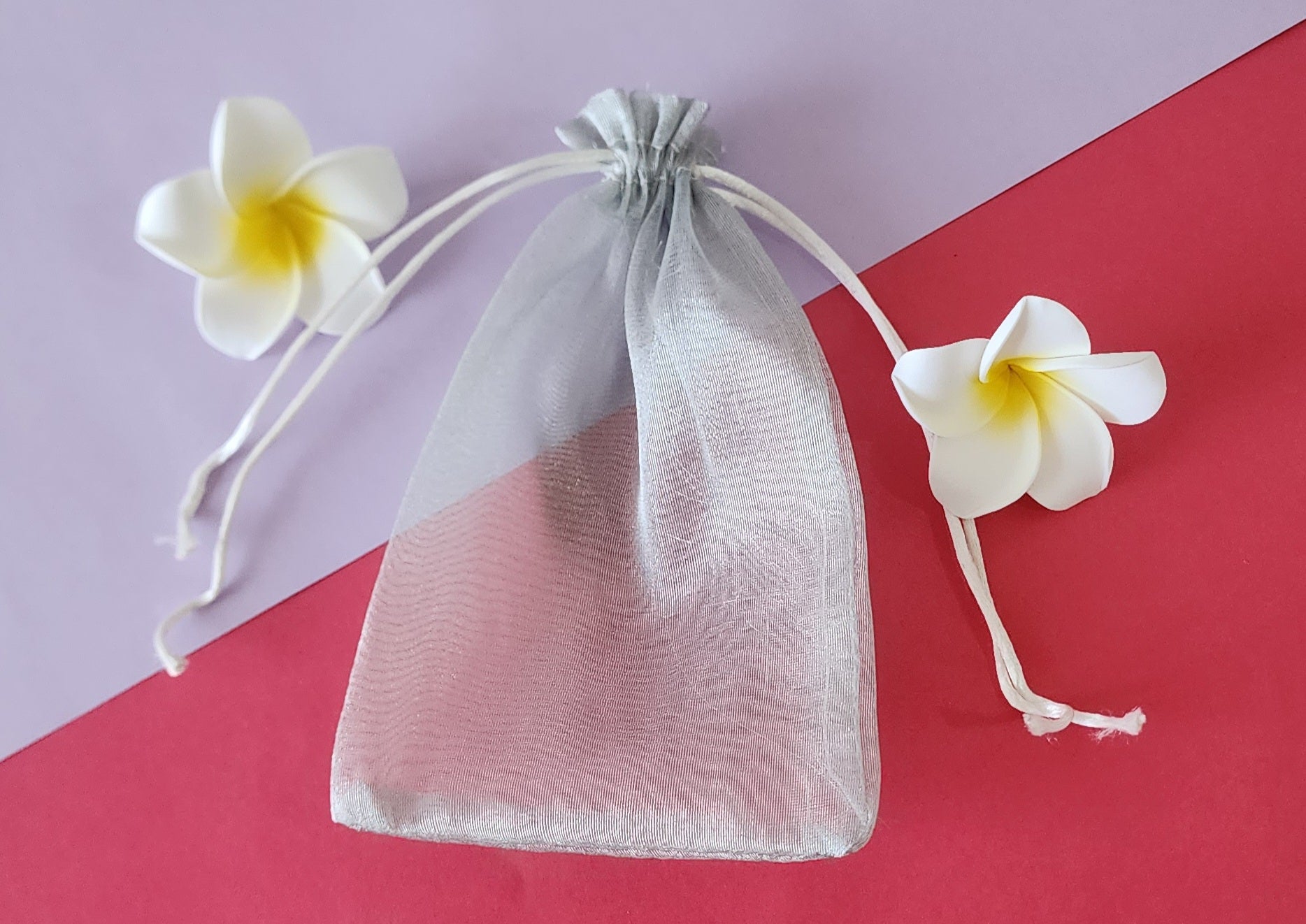 A clear, mesh-like fabric potli pouch with a drawstring closure, displayed on a red surface with two white flowers beside it.