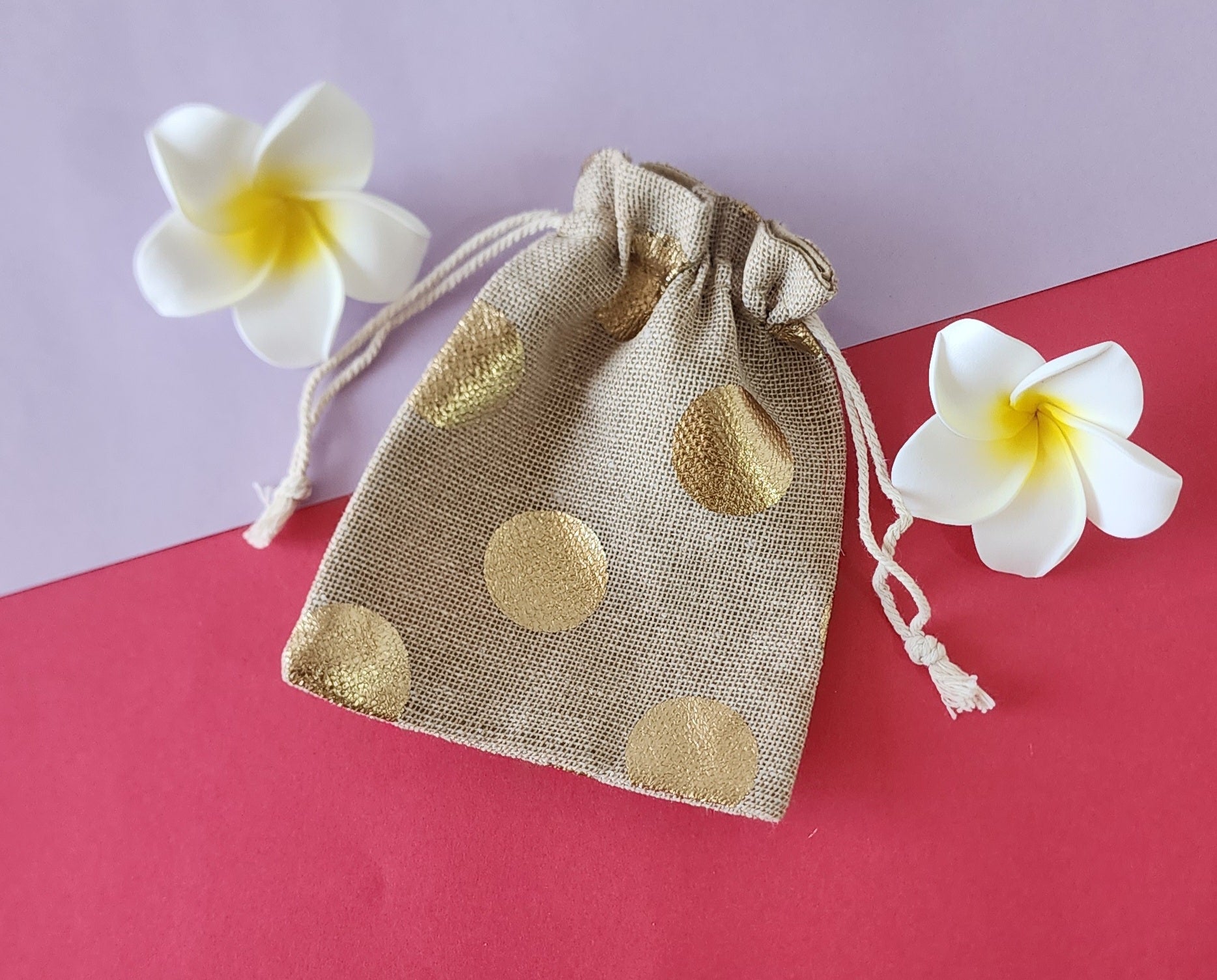 A red and gold patterned fabric potli pouch with a drawstring closure, accompanied by two white flowers on a green background.