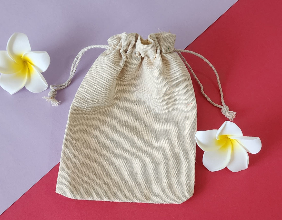 A beige Potli gift bag with a drawstring closure, displayed on a red surface with two white flowers beside it.
