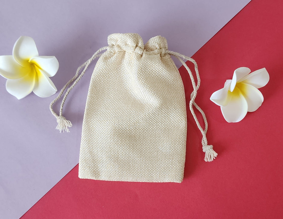 A beige Potli gift bag with a drawstring closure, displayed on a red surface with two white flowers beside it.