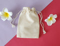 A beige Potli gift bag with a drawstring closure, displayed on a red surface with two white flowers beside it.
