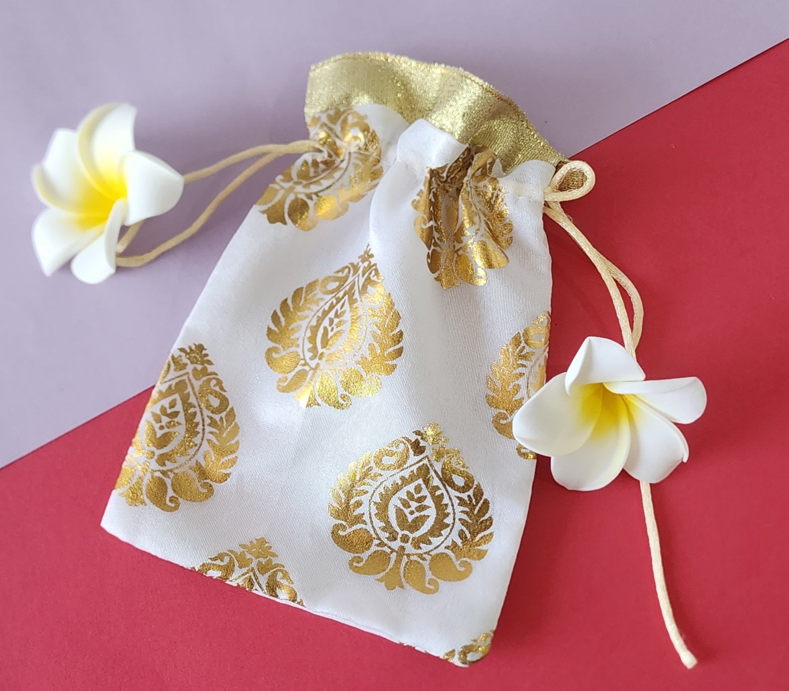 A red and gold patterned fabric potli pouch with a drawstring closure, accompanied by two white flowers on a green background.