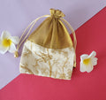 A gold patterned fabric potli pouch with a drawstring closure, accompanied by two white flowers on a green background.