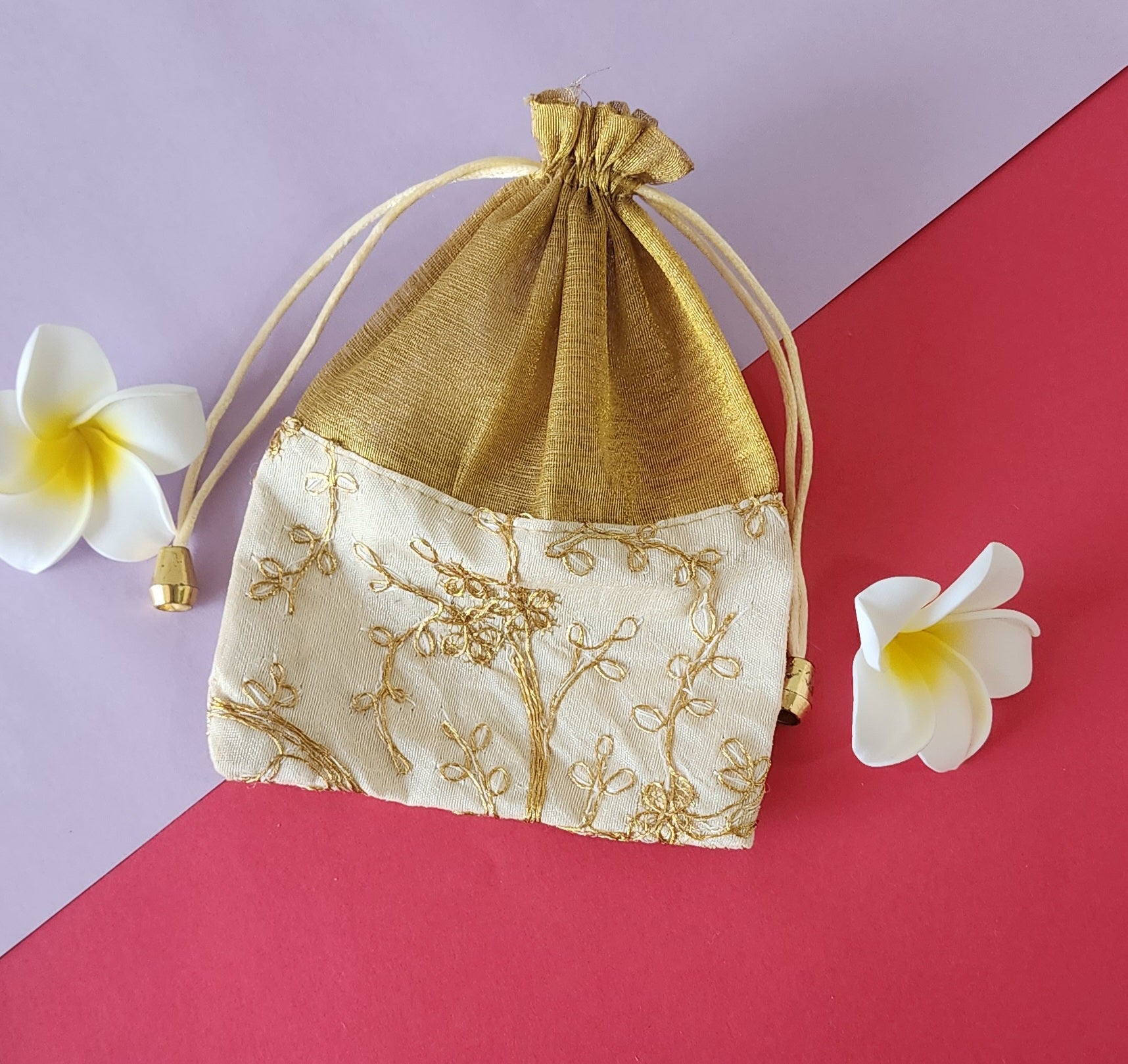 A gold patterned fabric potli pouch with a drawstring closure, accompanied by two white flowers on a green background.