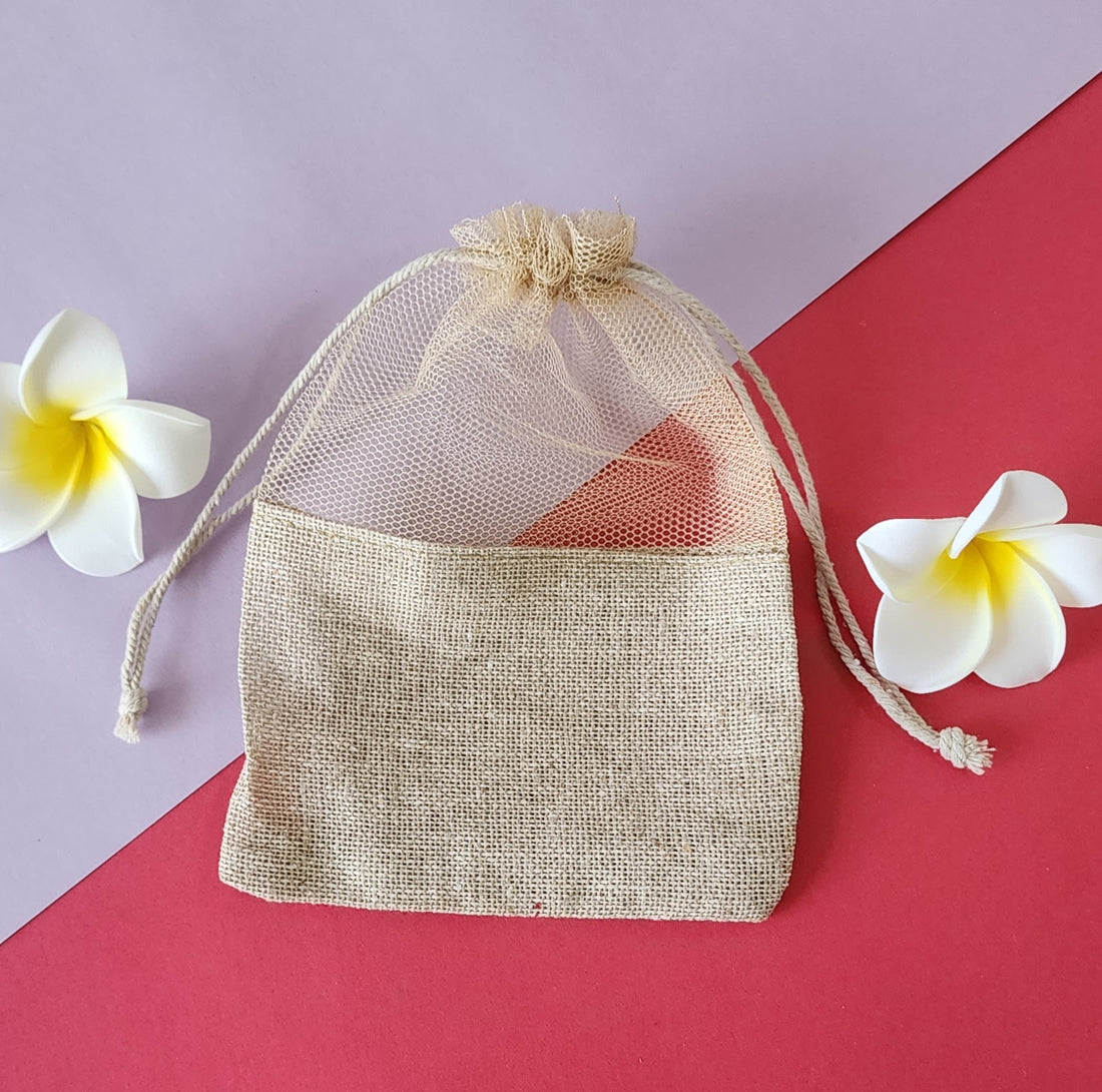 A clear, mesh-like fabric potli pouch with a drawstring closure, displayed on a red surface with two white flowers beside it.