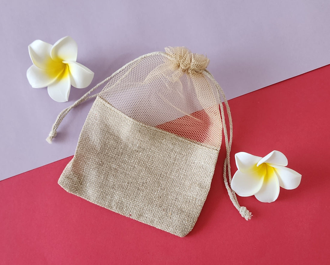 A clear, mesh-like fabric potli pouch with a drawstring closure, displayed on a red surface with two white flowers beside it.