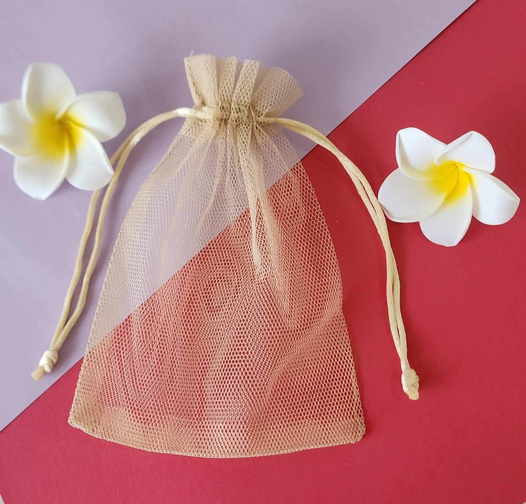 A clear, mesh-like fabric potli pouch with a drawstring closure, displayed on a red surface with two white flowers beside it.