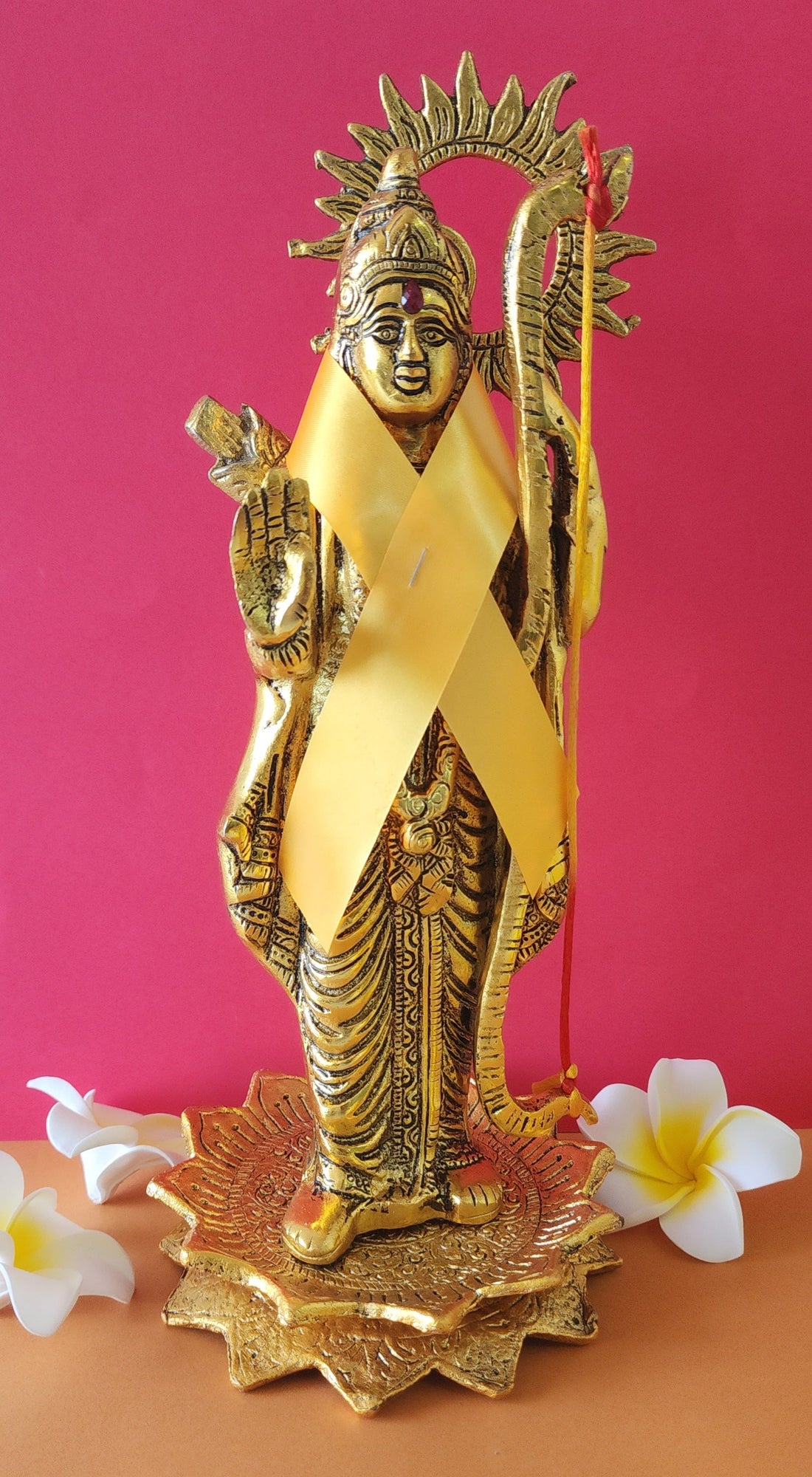 A golden metal statue of Lord Shri Ram stands against a vibrant pink background, adorned with a bright yellow ribbon and holding a golden bow and arrow, on a decorative base with white and yellow frangipani flowers at its feet.