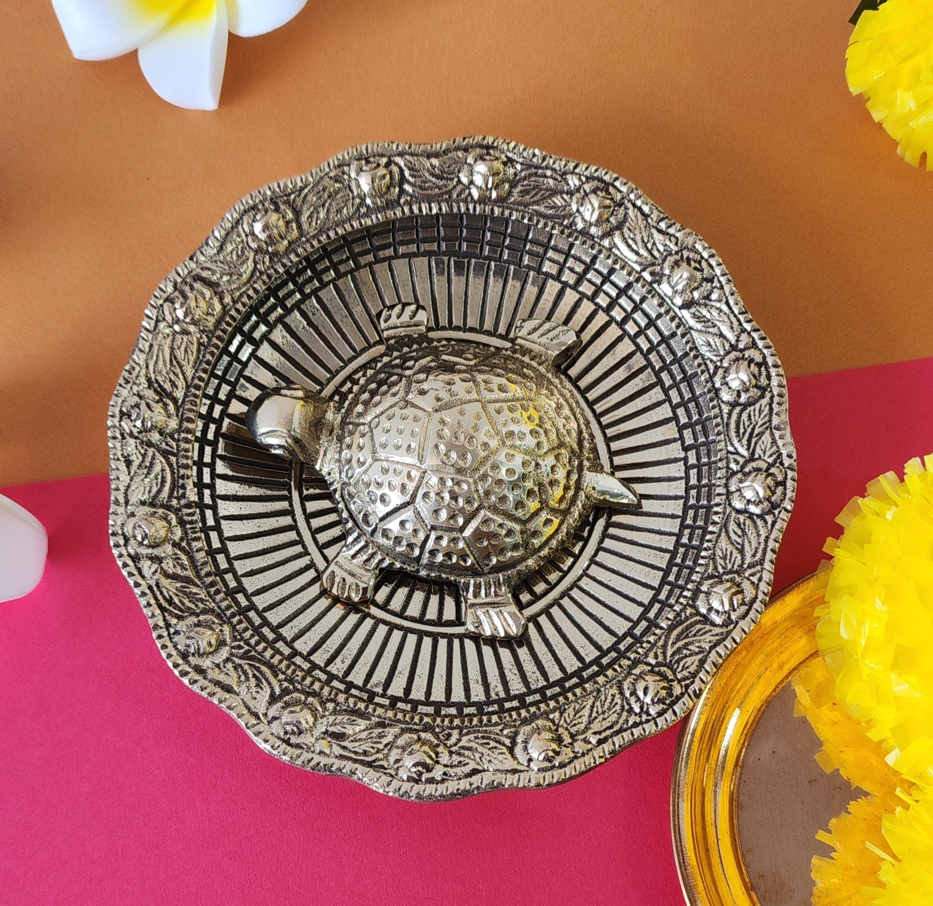 A metal turtle placed on a decorative plate with floral patterns, possibly used for Vaastu Fengshui purposes, displayed against a colorful background.
