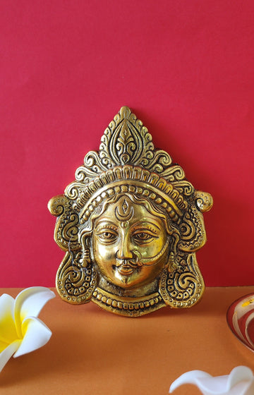 Golden face sculpture of Goddess Durga with intricate crown detail, displayed against a vibrant red and orange background with flowers.