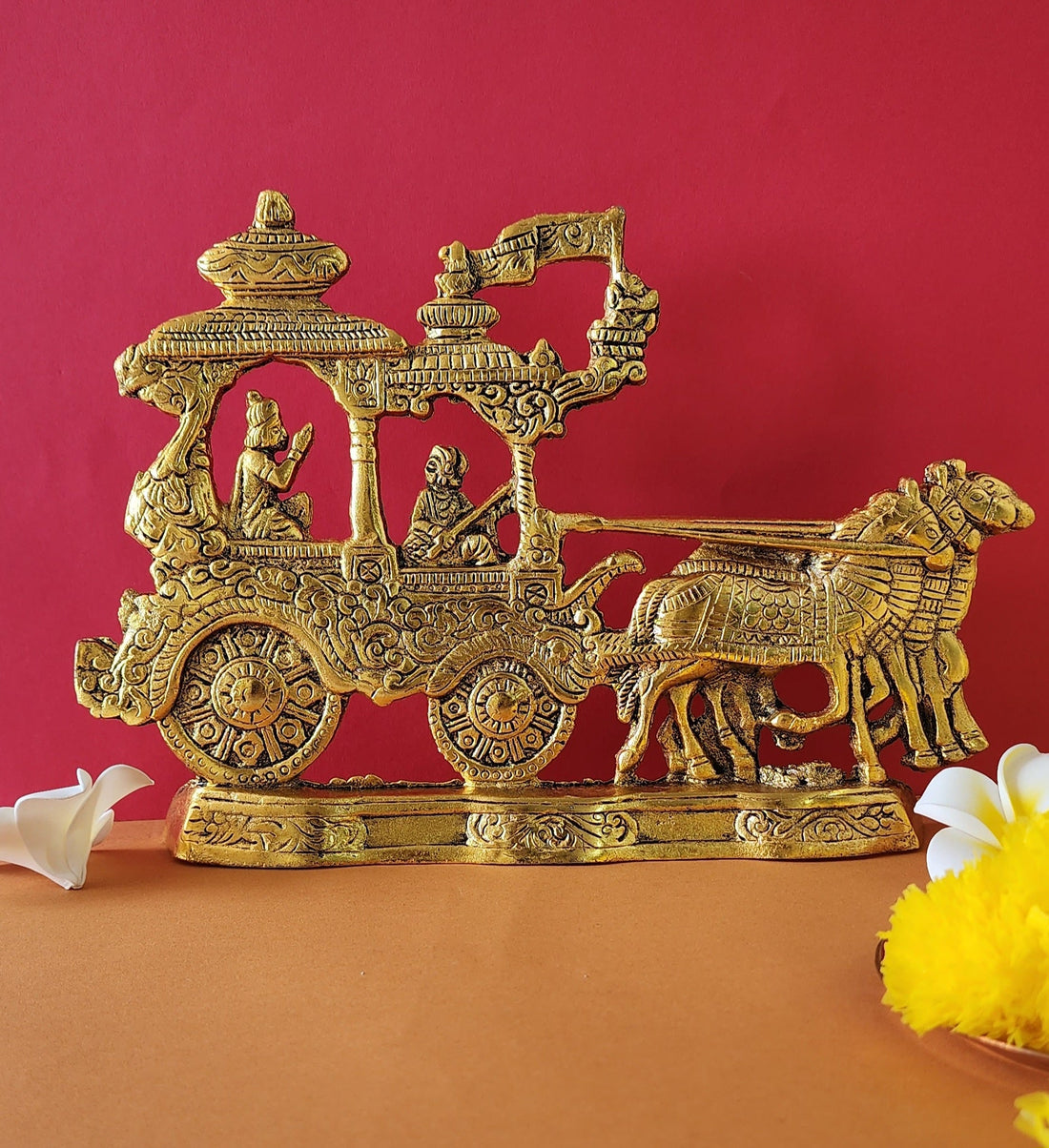A golden brass idol depicting the Mahabharata scene of Lord Krishna and Arjuna on their chariot (rath), pulled by four horses, with Lord Hanuman on the chariot's flag, all set against a vibrant reddish-orange background.