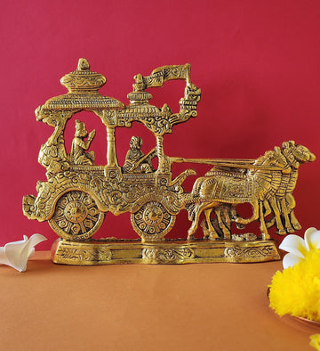 A golden brass idol depicting the Mahabharata scene of Lord Krishna and Arjuna on their chariot (rath), pulled by four horses, with Lord Hanuman on the chariot's flag, all set against a vibrant reddish-orange background.