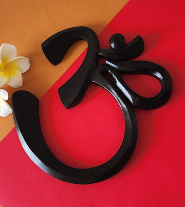 A black decorative wall art in the shape of the sacred symbol OM placed on a red and orange background, with a white flower to the side.