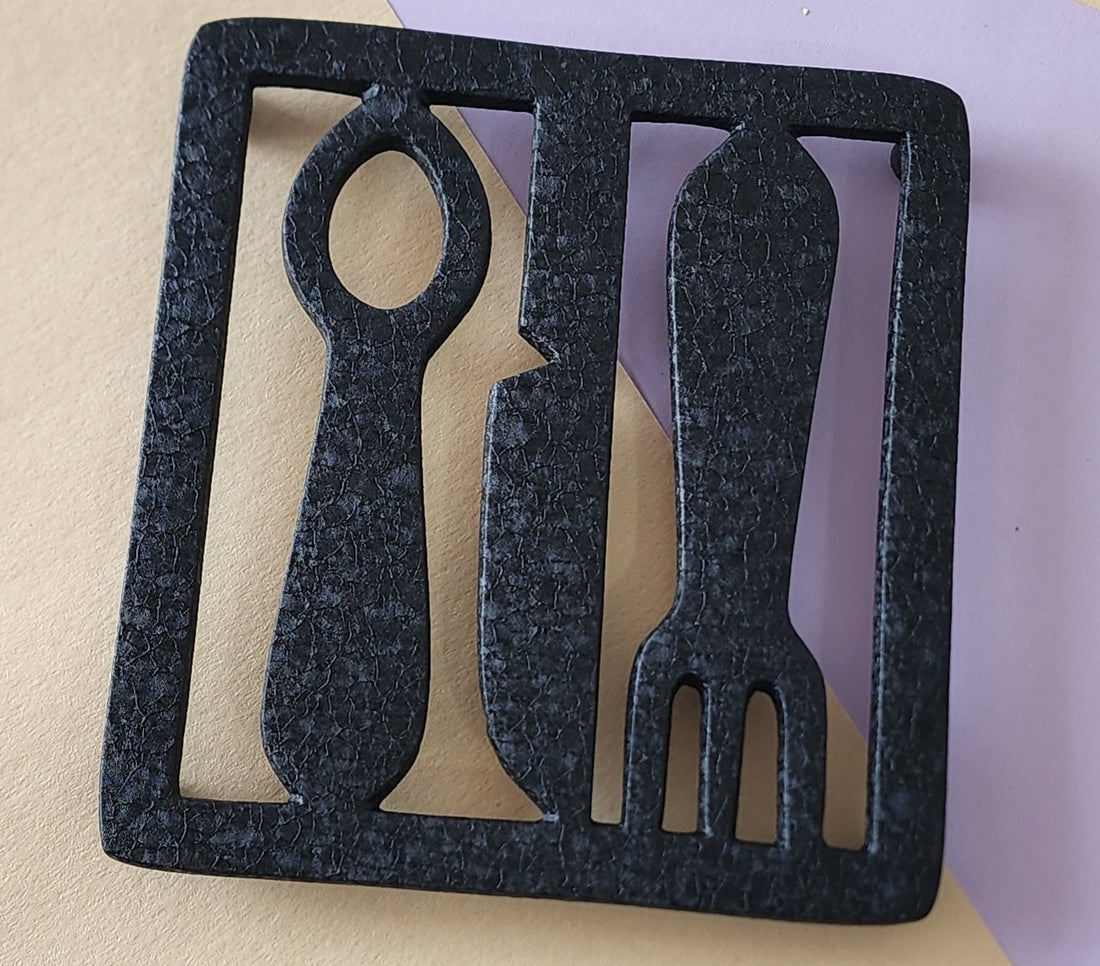 A black aluminum trivet designed with a fork and knife silhouette.