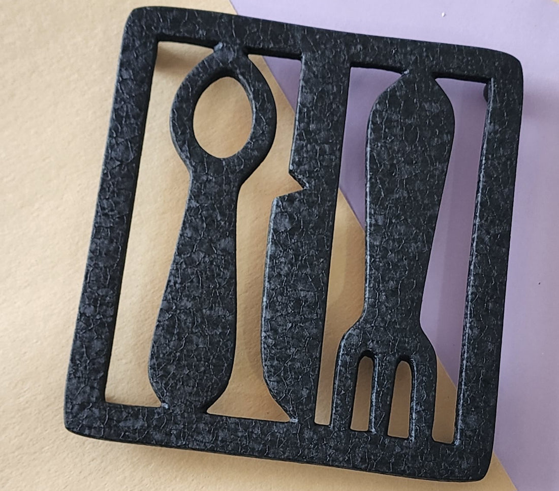 A black aluminum trivet designed with a fork and knife silhouette.