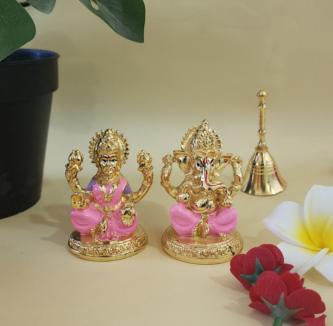 Gold and pink idols of Lakshmi and Ganesha on a shelf, with a small brass bell and flowers nearby.