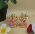 Golden brass idols of Goddess Lakshmi and Lord Ganesha, seated, with pink accents on their attire, placed indoors next to green foliage and flowers.