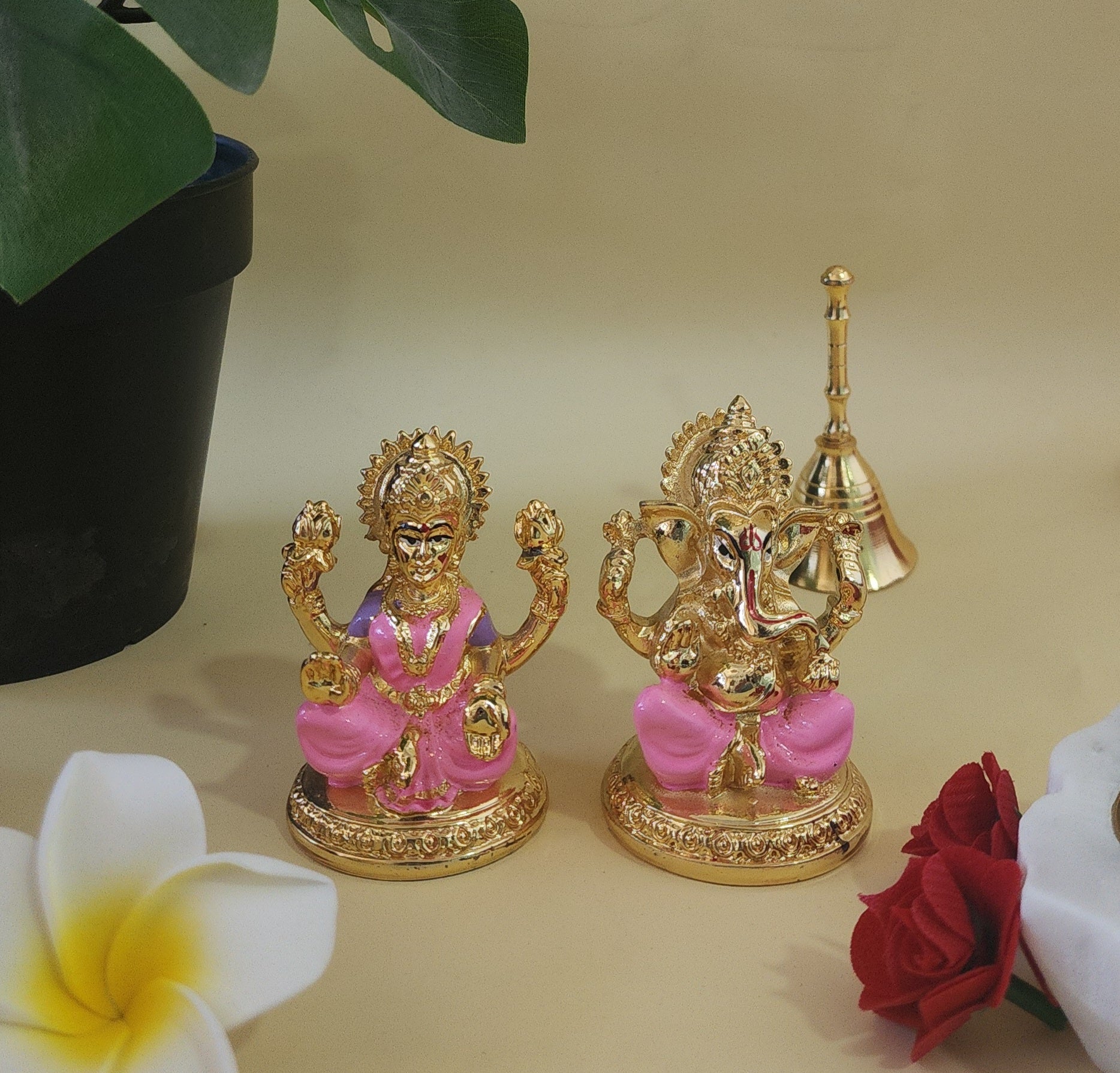 Golden brass idols of Goddess Lakshmi and Lord Ganesha, seated, with pink accents on their attire, placed indoors next to green foliage and flowers.