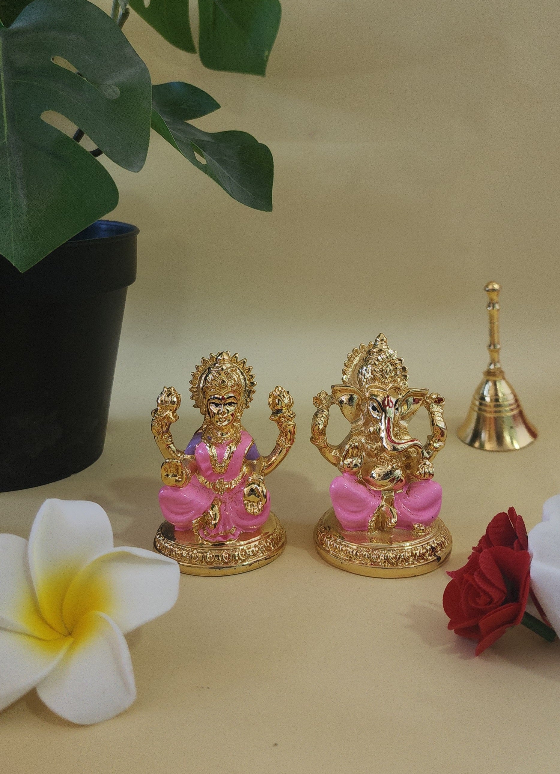 A gold-colored Lakshmi Ganesha idol set on a pink base, placed on a light-colored surface with a green plant in a black pot, a white flower, and red flowers in the background.