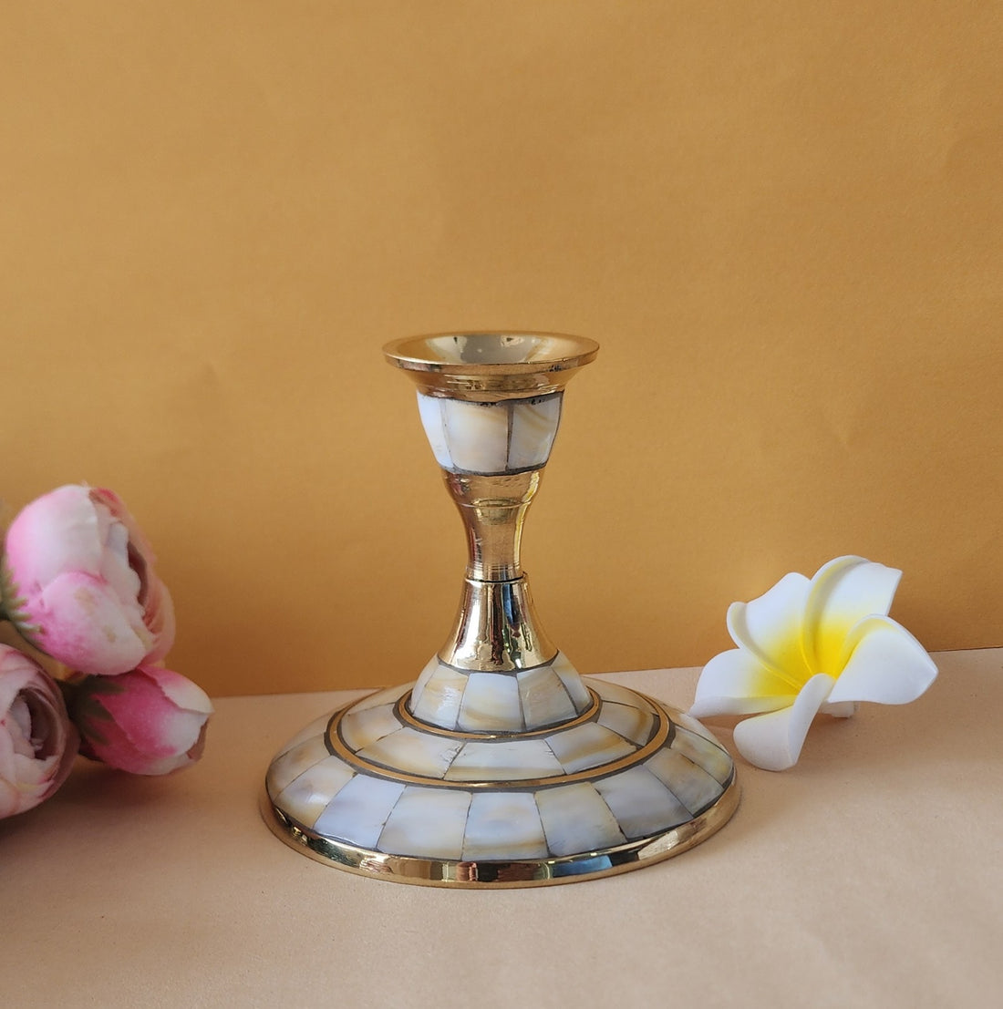 A single candle stand with a wide, circular base made of brass and inlaid with mother-of-pearl or similar iridescent material. The stand has a slender, tapering stem and is shown on a light-colored surface with two flowers on either side.